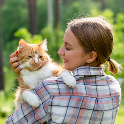 A girl with blonde hair in a blue, white, pink, and black plaid shirt holding her orange and white cat outside. In the background, there is a blurred forest line.