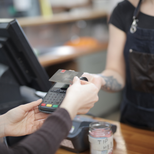 Cashier assisting a customer with a card payment at checkout. A ‘Tip me!’ jar is visible on the counter. The cashier, wearing an apron and with a tattoo on their arm, is slightly blurred.